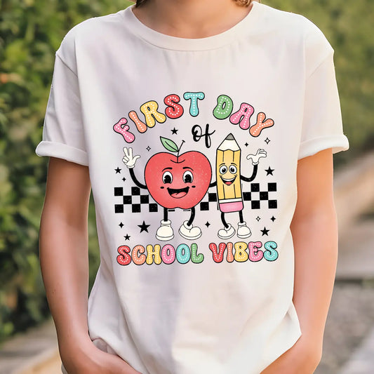 White T-shirt with “First Day of School Vibes” DTF transfer design featuring cute cartoon apple and pencil characters on a black checkerboard background.