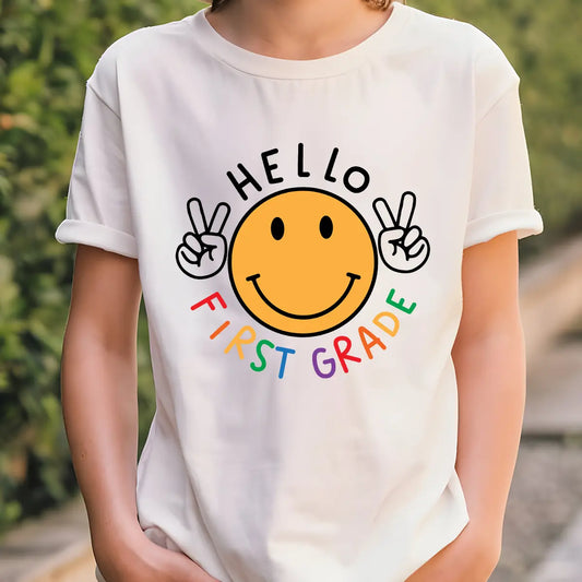 White T-shirt with “Hello First Grade” DTF transfer design featuring a big yellow smiley face with peace signs and colorful rainbow text.