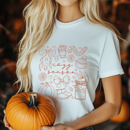 White T-shirt with “Cozy Season” DTF transfer design featuring autumn line-art icons like bows, pumpkin, scarf, coffee, boots, candle, and flowers.