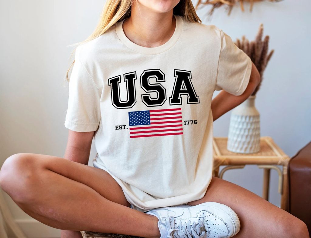 Cream-colored t-shirt with bold USA varsity letters, an American flag, and EST. 1776 text, perfect for 4th of July Ready-to-Press DTF Transfers.