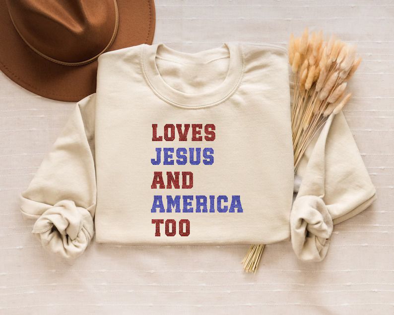 Cream-colored sweatshirt with Loves Jesus and America Too text in red, white, and blue distressed font, ideal for Christian 4th of July DTF transfers.