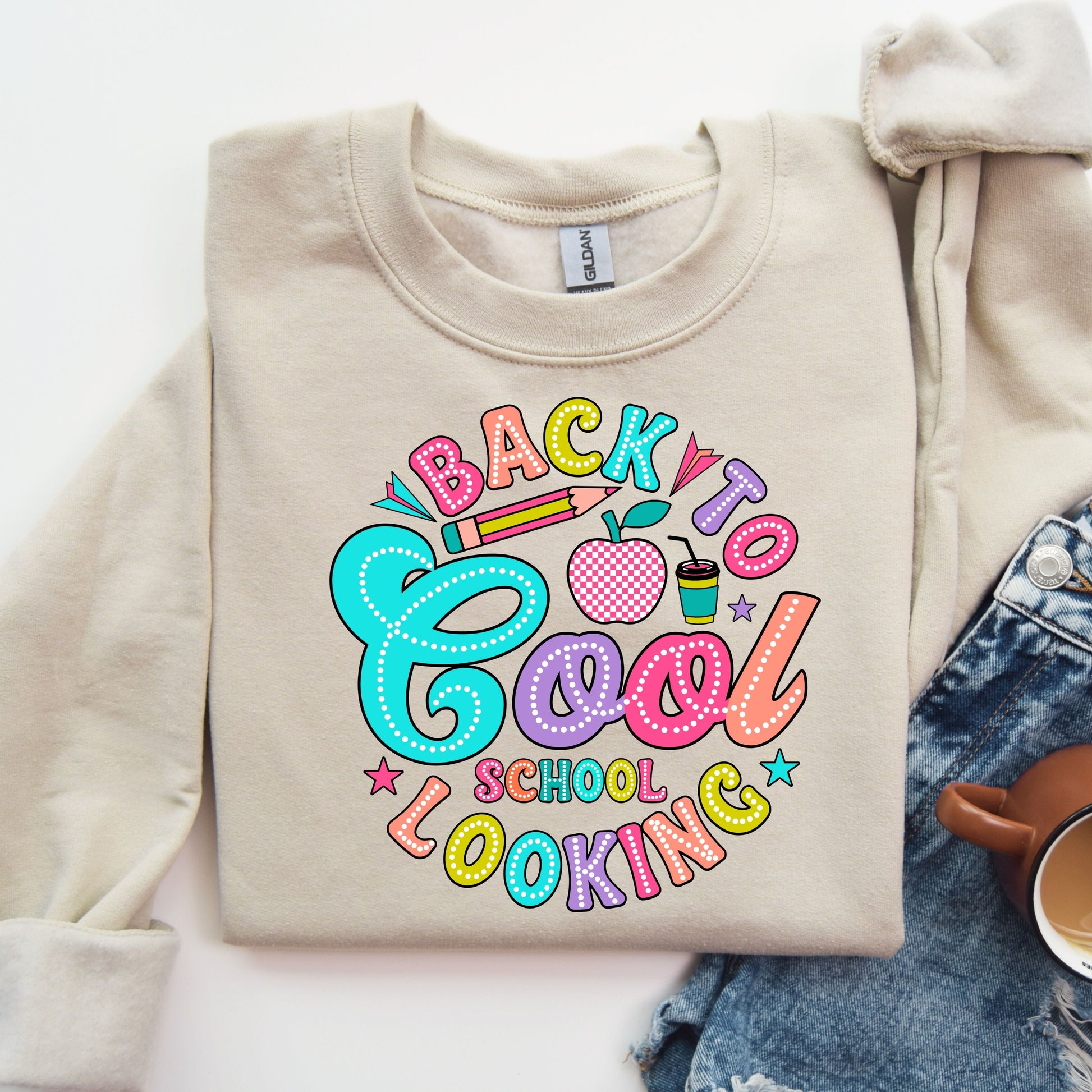 "Back to Cool School Looking" DTF transfer design on a beige sweatshirt featuring colorful, bold letters with school-themed graphics like pencils, an apple, and a drink cup.