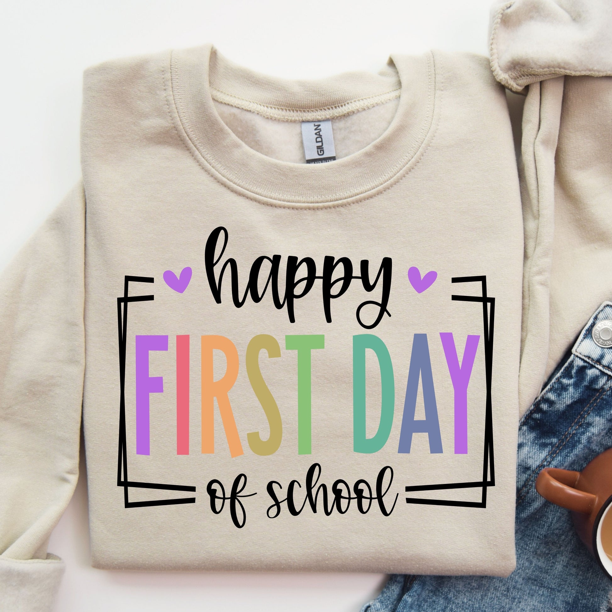 "Happy First Day of School" DTF transfer design on a beige sweatshirt featuring colorful pastel letters with heart accents and modern framing, perfect for back-to-school apparel.