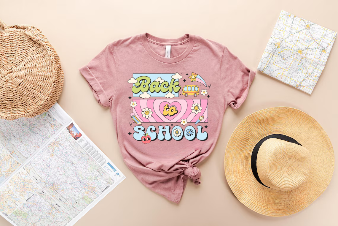 "Back to School" Retro-Themed DTF transfer design on a pink t-shirt featuring colorful retro fonts, daisies, clouds, a school bus, pencils, and apples, perfect for back-to-school apparel.