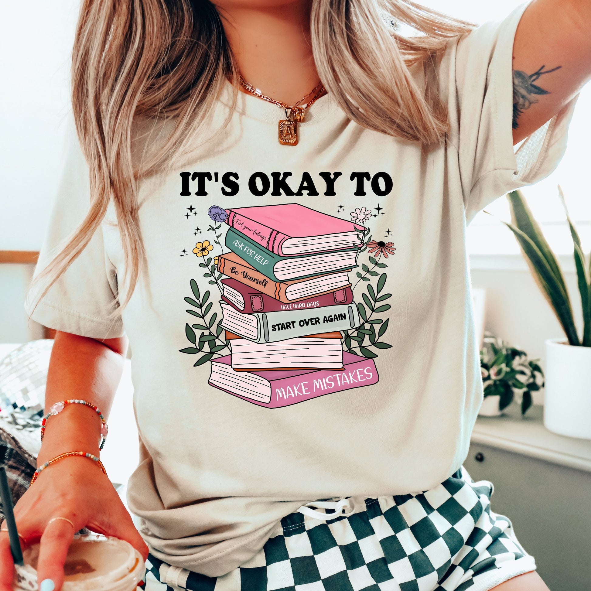 "It's Okay to Make Mistakes" DTF transfer design on a light beige t-shirt featuring a colorful stack of books with motivational messages, surrounded by flowers and greenery.