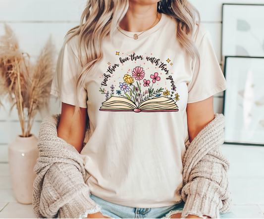 Beige t-shirt with an open book design, flowers growing from pages, and "Teach Them Love Them Watch Them Grow" text.