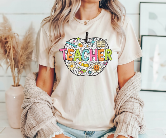 Beige t-shirt with a colorful apple-shaped design featuring the word “TEACHER” with school-themed elements like pencils, books, and stars.