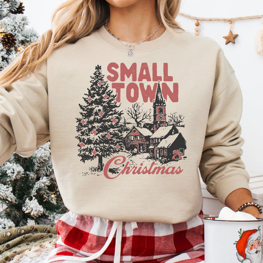 A charming small town Christmas scene on a beige sweatshirt, complete with a church, snow-covered trees, and rustic homes.