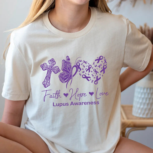 Beige t-shirt featuring a purple Faith Hope Love Lupus Awareness DTF design with a cross, butterfly, and heart made of ribbons.

