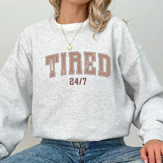Woman wearing a gray sweatshirt with bold brown varsity-style "Tired 24/7" text, featuring DTF Transfers ready to press for funny custom mom-life apparel.