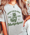 Woman wearing gray tee with “Mind Your Own Motherhood” text and yoga raccoon artwork—featuring DTF Transfers ready to press for funny mom-themed designs.