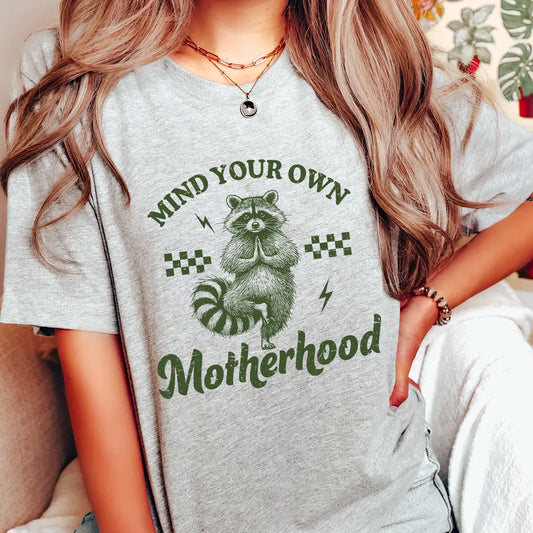 Woman wearing gray tee with “Mind Your Own Motherhood” text and yoga raccoon artwork—featuring DTF Transfers ready to press for funny mom-themed designs.