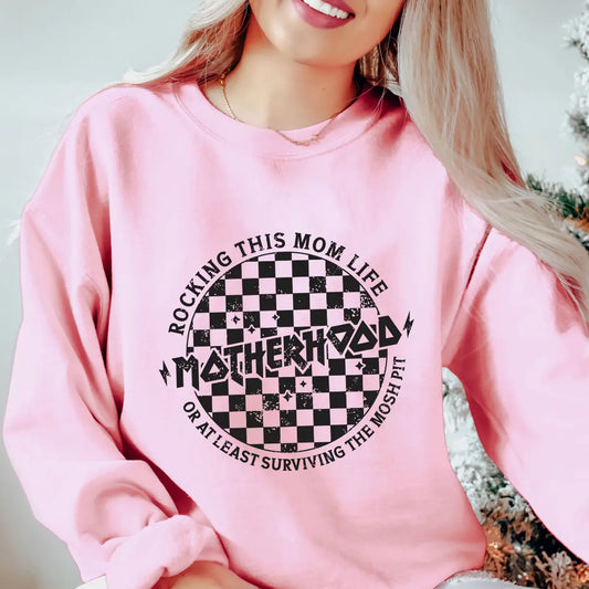 DTF Transfers ready to press with checkered grunge design and “Motherhood – Rocking This Mom Life or At Least Surviving the Mosh Pit” text on a pink sweatshirt.