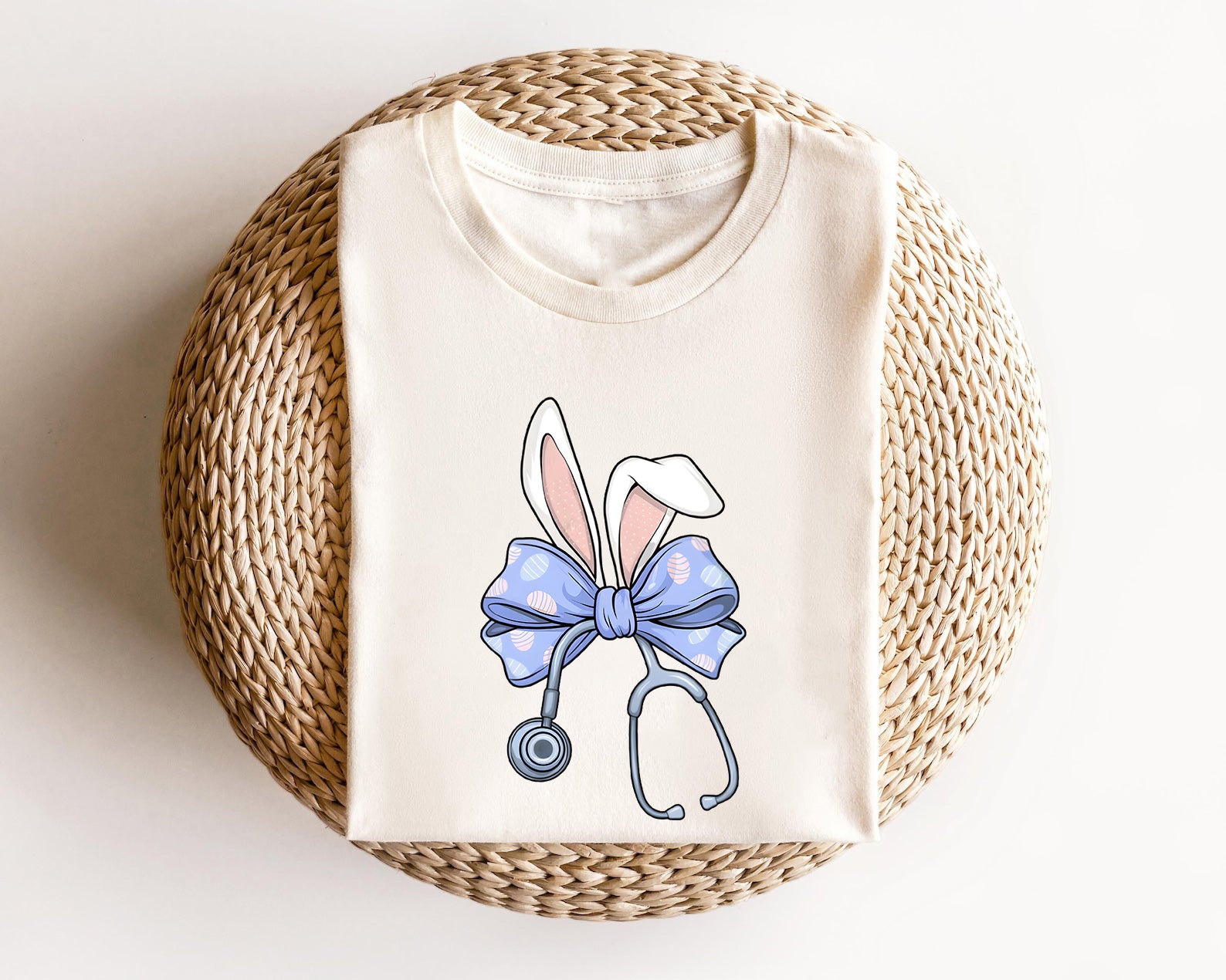 Cream-colored t-shirt with a cute Easter Bunny Nurse design featuring bunny ears, a stethoscope, and a pastel blue bow with Easter egg patterns.