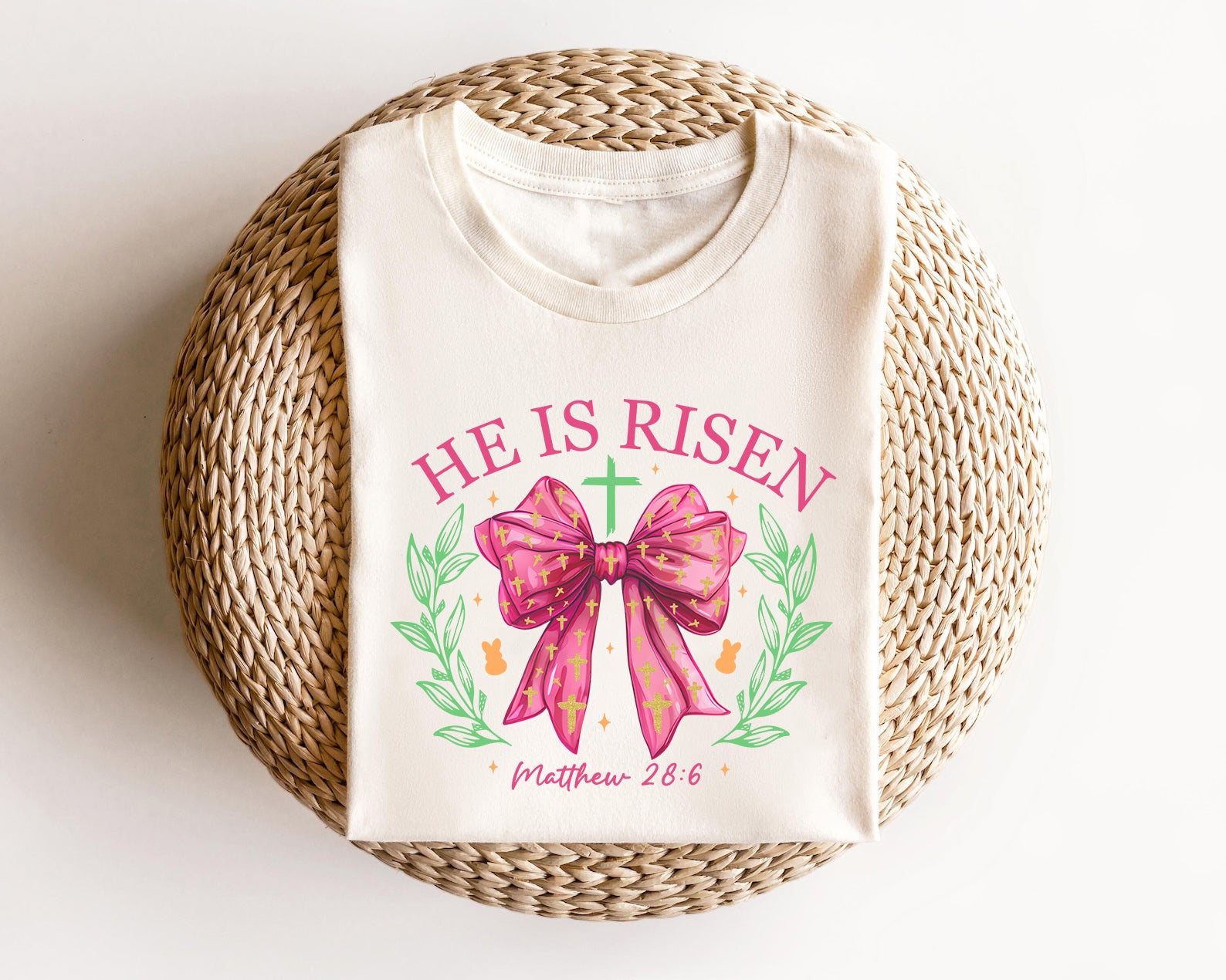 Cream-colored t-shirt featuring a "He Is Risen" design with a pink bow, golden crosses, and a green cross, perfect for Christian Easter celebrations.