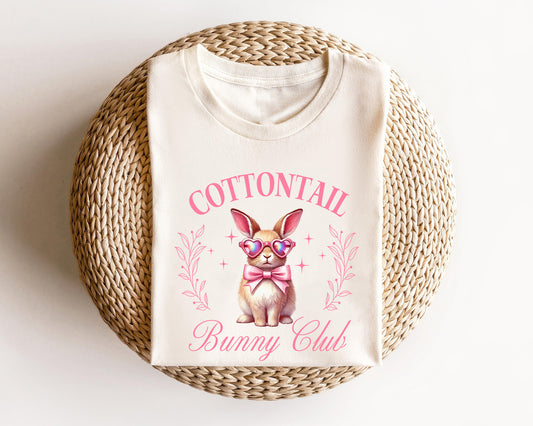 Cream-colored t-shirt featuring a cute bunny with heart sunglasses, a pink bow, and "Cottontail Bunny Club" text, perfect for Easter fashion.