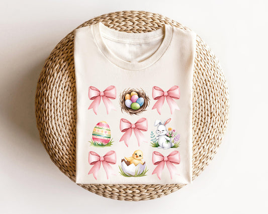 Cream-colored t-shirt featuring pastel Easter elements, including a bunny, egg nest, chick, decorated egg, and pink bows, perfect for Easter fashion.