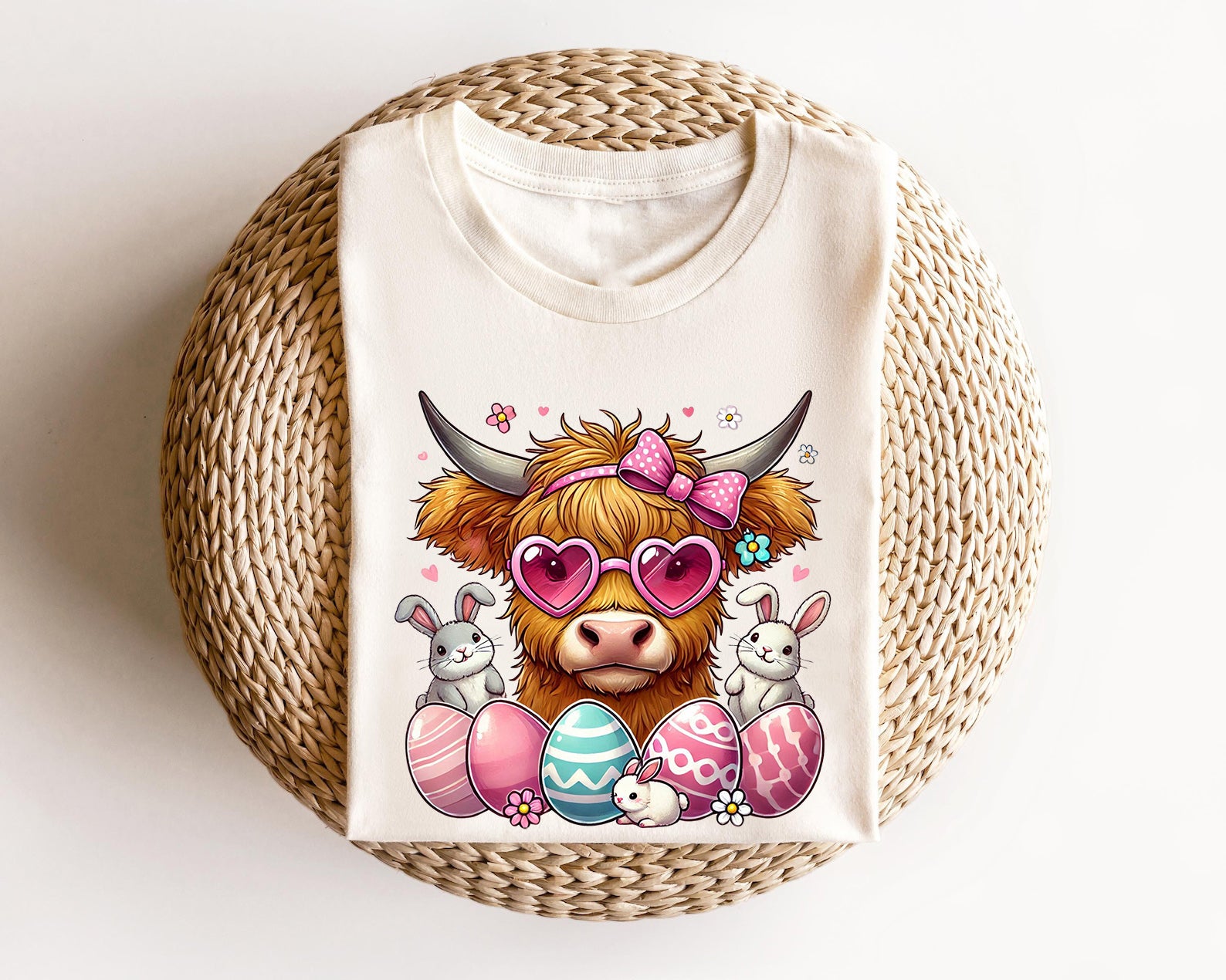 Cream-colored t-shirt featuring a cute highland cow with pink sunglasses, a bow, Easter eggs, and bunnies, perfect for Easter farm-style fashion.

