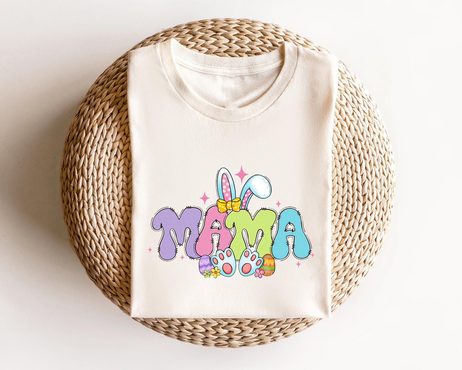 Cream-colored t-shirt featuring "MAMA" text with bunny ears, pastel colors, and Easter-themed elements, perfect for moms celebrating Easter.