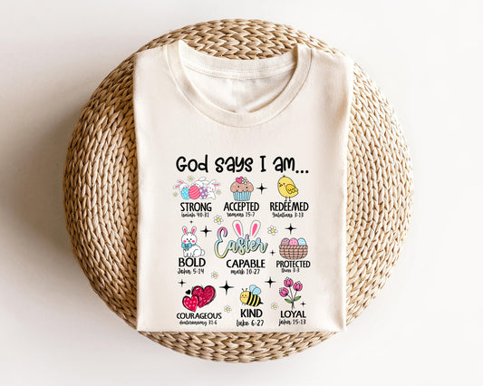 Cream-colored t-shirt featuring "God Says I Am" Christian Easter design with Bible verses, pastel Easter icons, and uplifting words of faith.