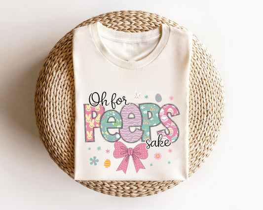 Cream-colored t-shirt featuring a colorful "Oh for Peeps Sake" text design with bunnies, Easter eggs, and a pink bow, perfect for Easter fun.