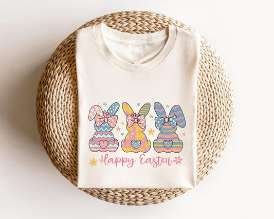 Cream-colored t-shirt featuring three pastel-patterned bunnies with bows, heart accents, and "Happy Easter" text, perfect for Easter celebrations.