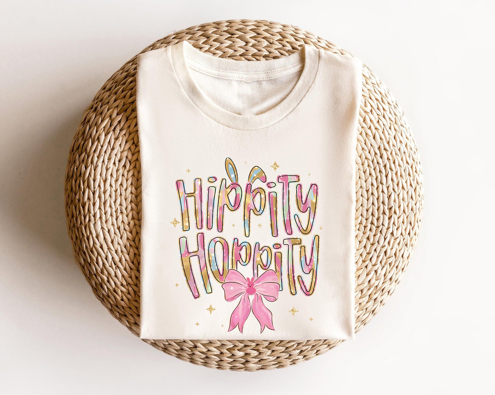 Cream-colored t-shirt featuring "Hippity Hoppity" text with bunny ears, pastel colors, sparkles, and a pink bow, perfect for Easter fun.