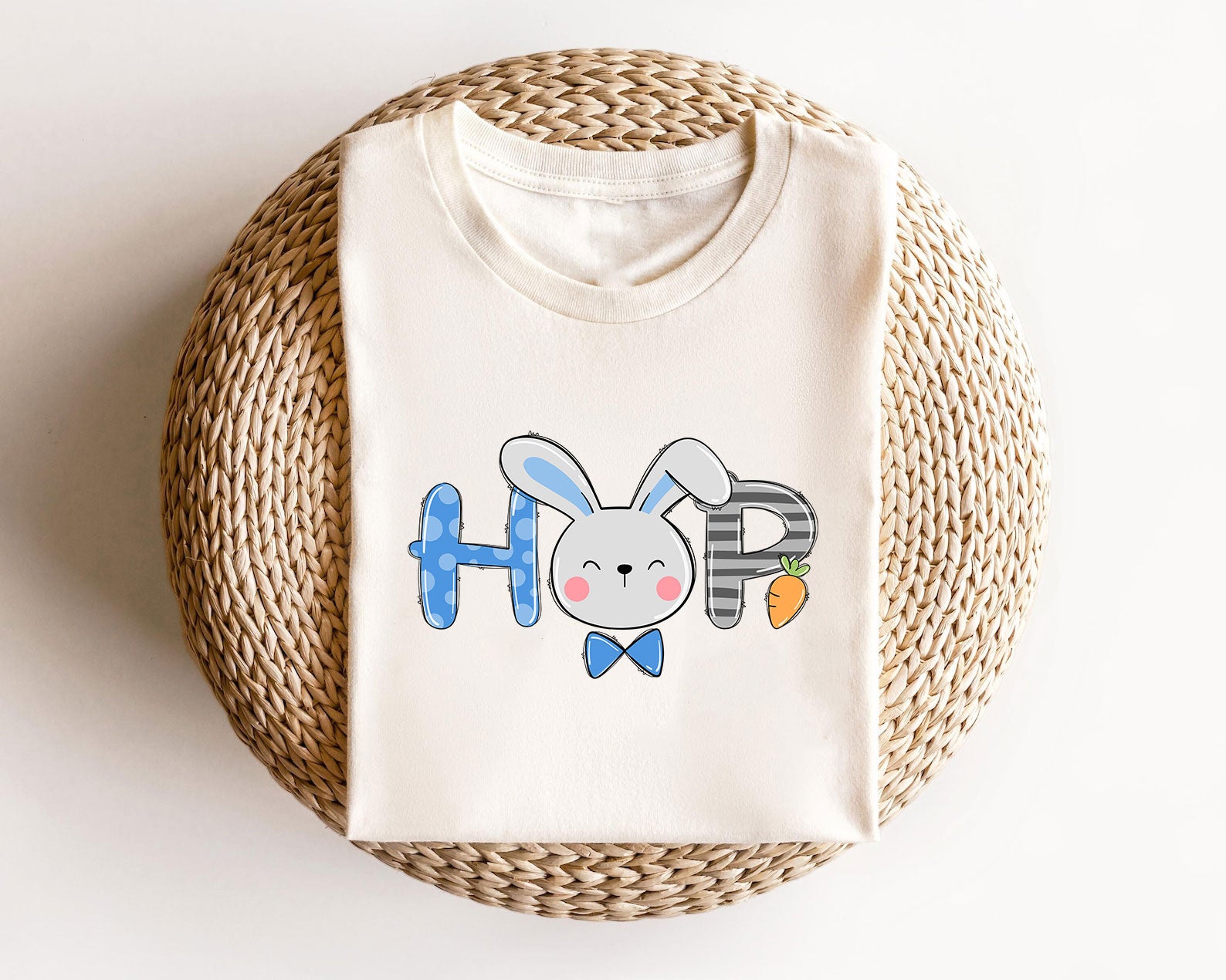 Cream-colored t-shirt with "HOP" text, featuring a cute bunny face as the letter "O," blue and plaid patterns, and a small carrot.