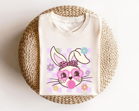 Cream t-shirt featuring a stylish bunny face with pink sunglasses, a cheetah bandana, and a pastel lilac checkerboard background—perfect for Easter apparel.