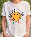 White T-shirt with “Hello First Grade” DTF transfer design featuring a big yellow smiley face with peace signs and colorful rainbow text.