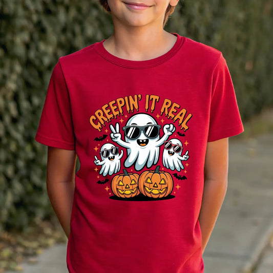 Red t-shirt with Halloween design featuring three cartoon ghosts in sunglasses, jack-o’-lantern pumpkins, bats, stars, and text “Creepin’ It Real.”