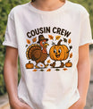 White t-shirt with Thanksgiving design featuring a cartoon turkey in a pilgrim hat and a smiling pumpkin surrounded by autumn leaves, with the text “Cousin Crew.”