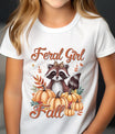 White t-shirt with autumn design featuring a raccoon with leopard bow surrounded by pumpkins and fall leaves, text “Feral Girl Fall” in script style.