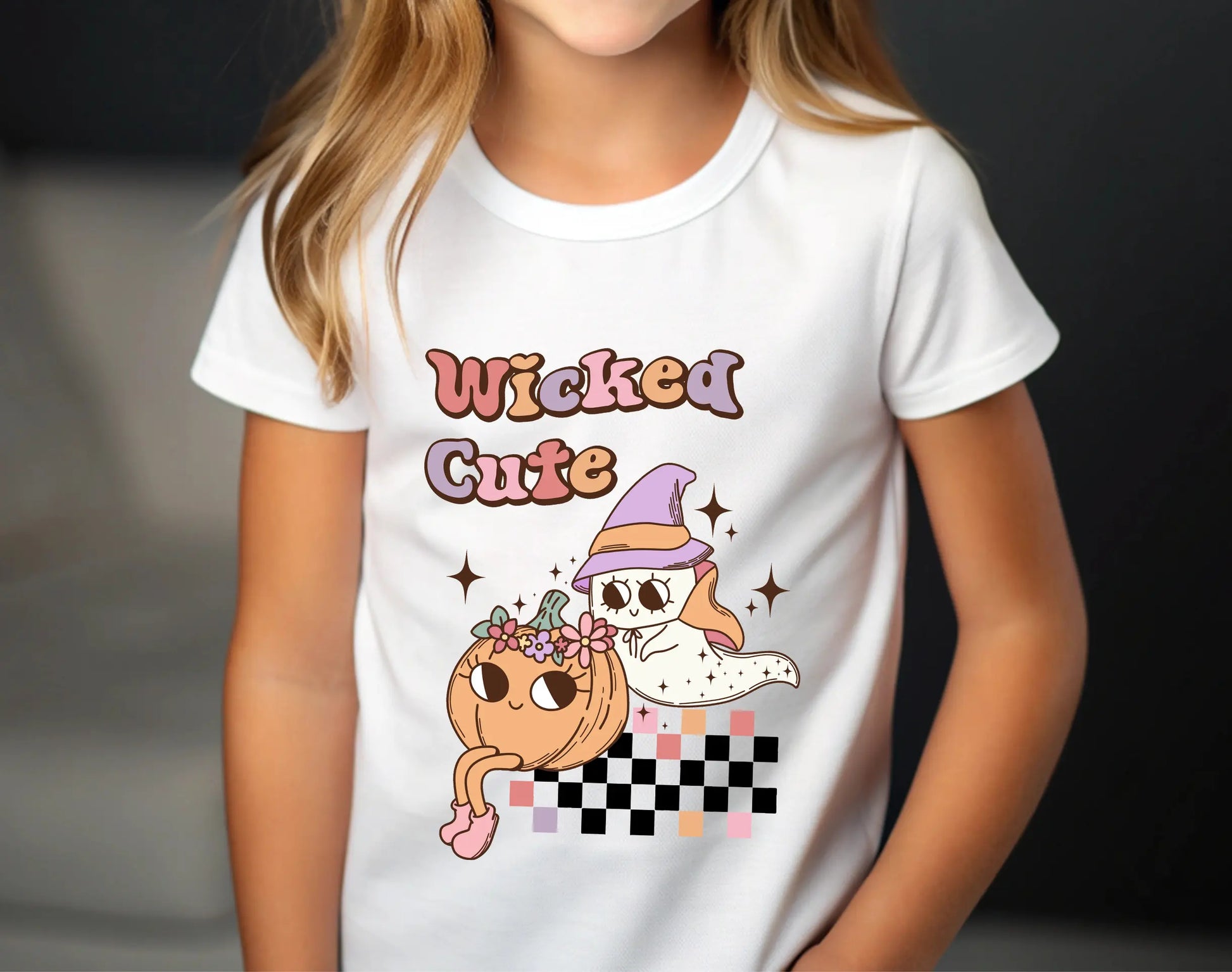 White t-shirt with Halloween design featuring a cute ghost in a pastel witch hat, smiling pumpkin with flowers, retro “Wicked Cute” text, and checkerboard accent.