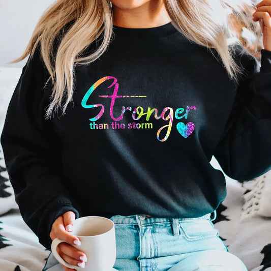 Black sweatshirt with rainbow gradient text “Stronger Than the Storm” and heart accent DTF transfer design, inspirational and motivational apparel.