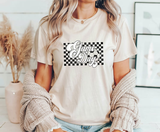 Woman wearing a beige t-shirt featuring "Game Day" in stylish script over a checkered soccer-themed background, perfect for soccer moms and players.