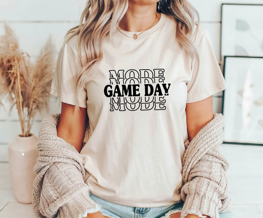 Woman wearing a beige t-shirt featuring bold stacked "MODE" text with "GAME DAY" in black, perfect for game day apparel.