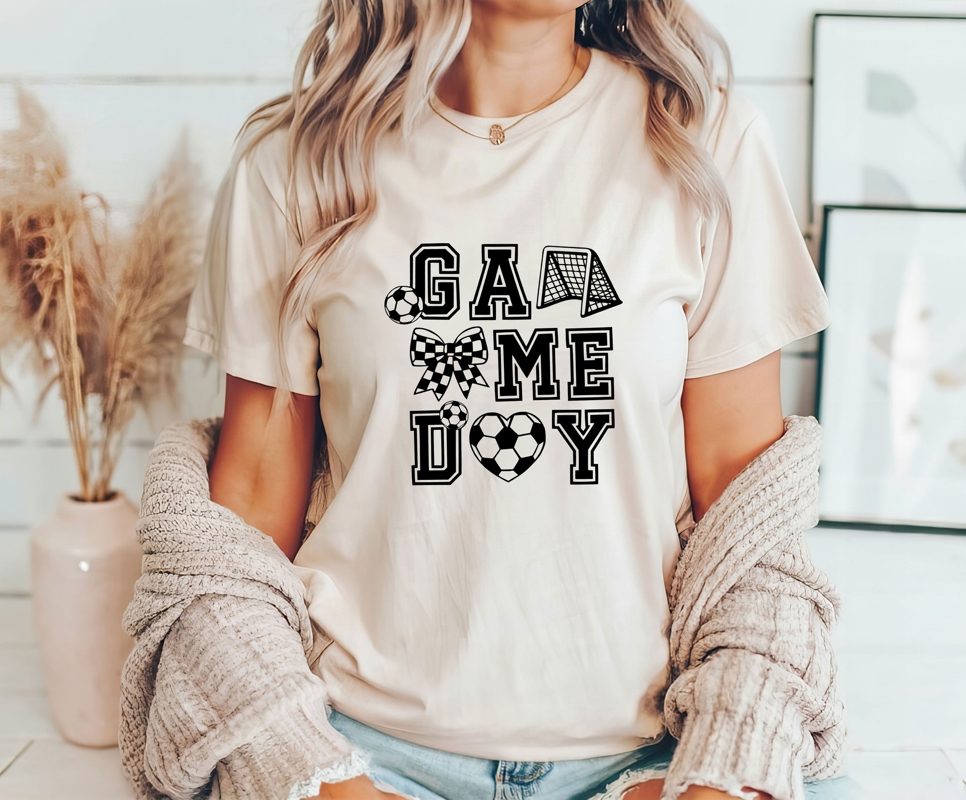 Woman wearing a beige t-shirt featuring "GAME DAY" text with soccer-themed elements, including a goal post, bow, soccer ball, and heart, perfect for soccer fans and moms.