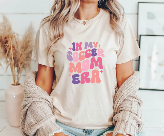 Woman wearing a beige t-shirt featuring "In My Soccer Mom Era" in colorful retro-style text, perfect for stylish soccer moms.