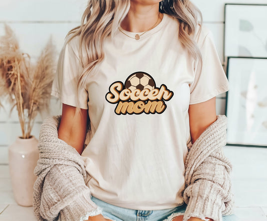 Woman wearing a beige t-shirt featuring "Soccer Mom" in a retro font with a soccer ball accent, perfect for stylish soccer moms.