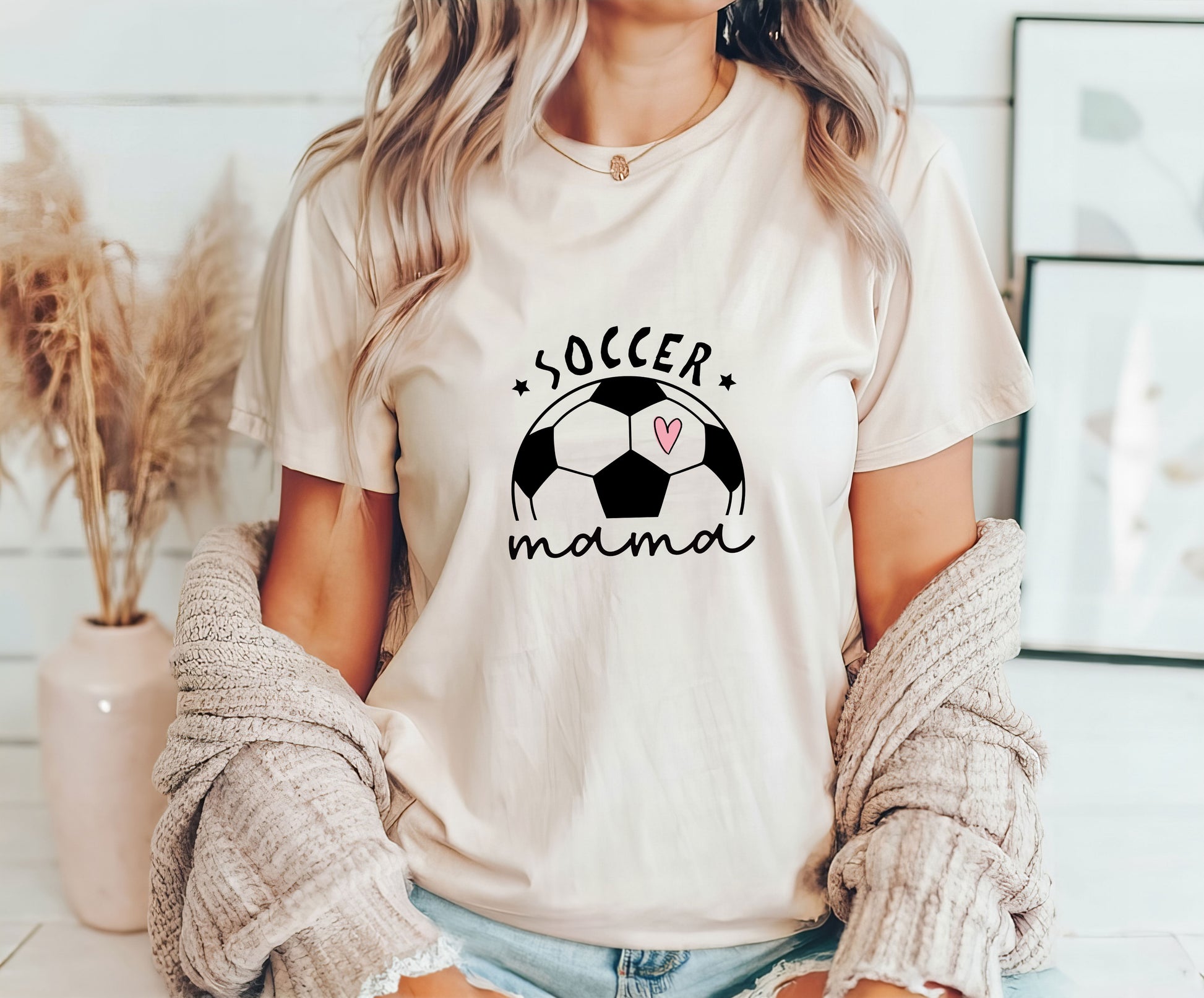 Woman wearing a beige t-shirt featuring a soccer ball with a pink heart and the words "Soccer Mama," perfect for stylish soccer moms.