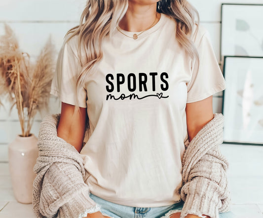 Woman wearing a beige t-shirt featuring "Sports Mom" in a minimalist font with a heart accent, perfect for stylish sports moms.