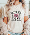 Woman wearing a beige t-shirt with a Pink Bow Soccer Mom Social Club design featuring a soccer ball, laurel wreath, and stylish typography.