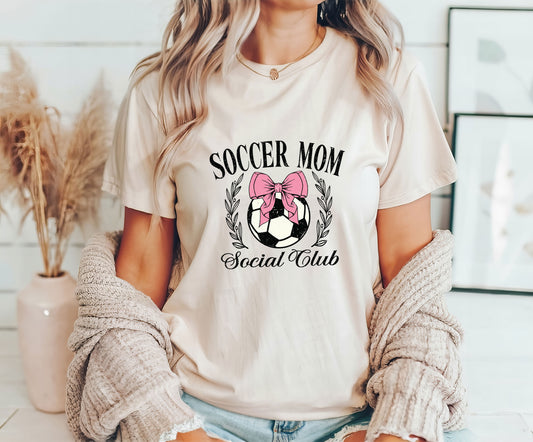 Woman wearing a beige t-shirt with a Pink Bow Soccer Mom Social Club design featuring a soccer ball, laurel wreath, and stylish typography.