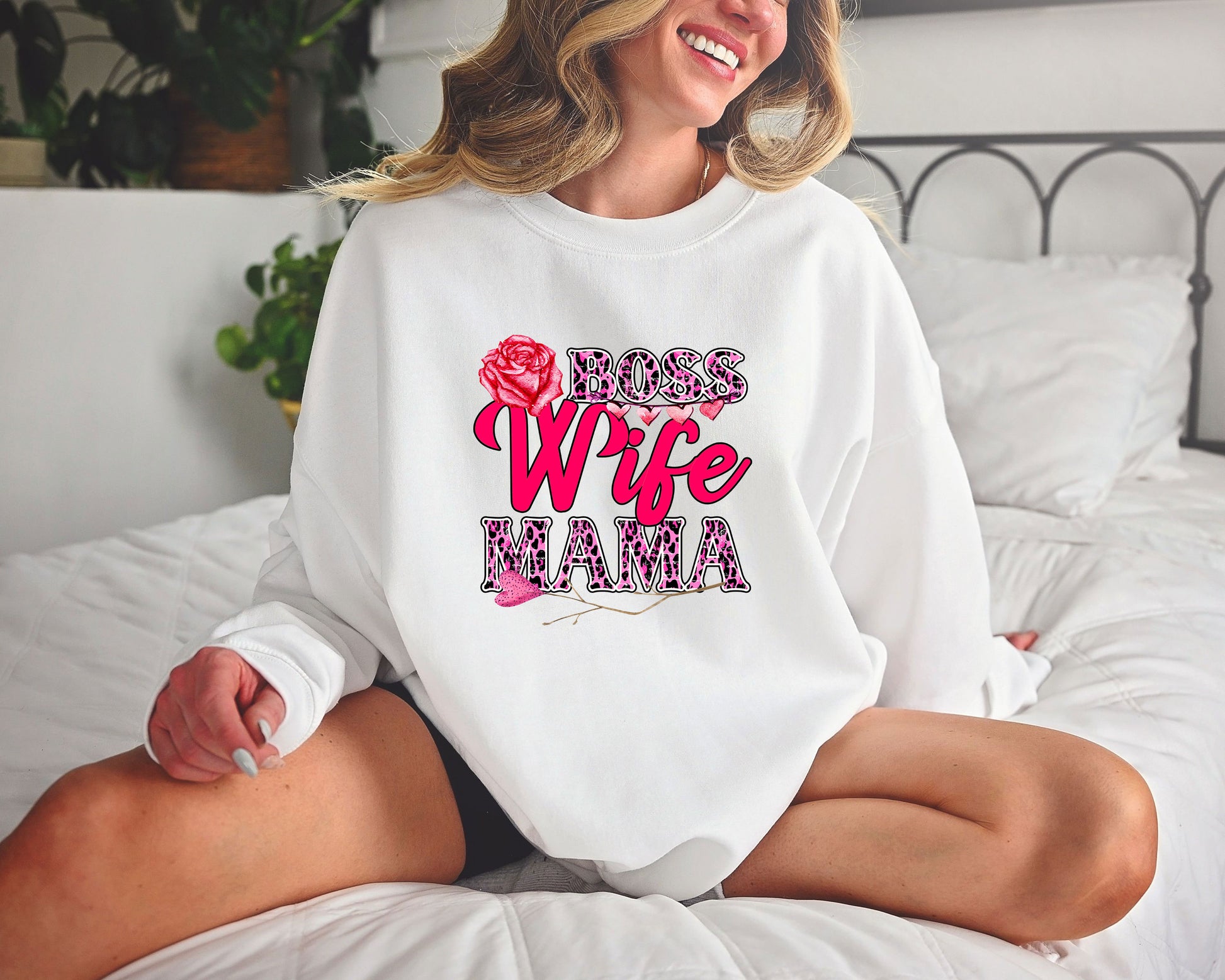 Woman wearing a white sweatshirt featuring a Boss Wife Mama design with pink and leopard print accents, a red rose, and stylish typography.