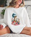 A woman wearing a white sweatshirt featuring a Hot Mess Mama DTF Transfer, depicting a skeleton mom sipping coffee with a towel wrap and floral accents.