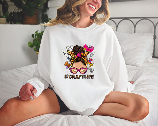 A woman wearing a white sweatshirt featuring a #CraftLife DTF Transfer, showcasing a messy bun hairstyle with crafting tools, perfect for DIY moms and crafters.