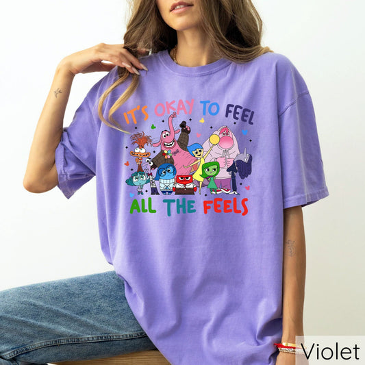 "It's Okay to Feel All the Feels" DTF transfer design on a purple t-shirt featuring a colorful group of characters and uplifting text.