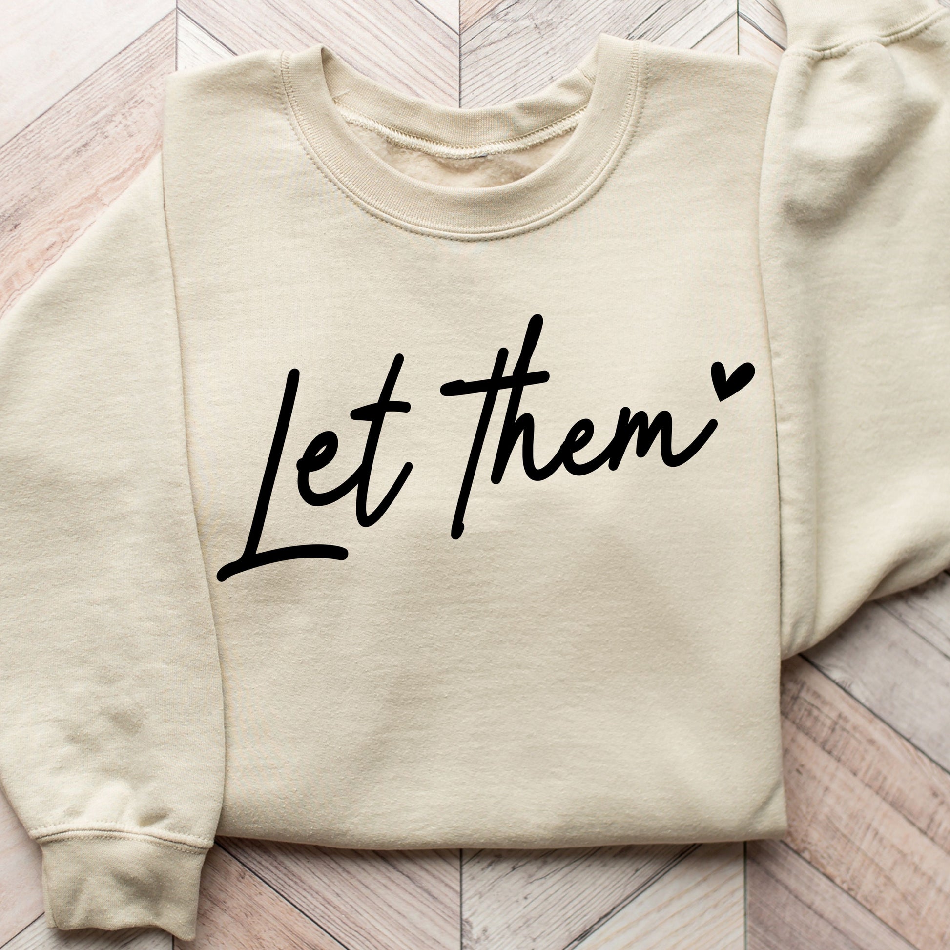 "Let Them" DTF transfer design on a beige sweatshirt featuring elegant script typography and a heart detail in black.