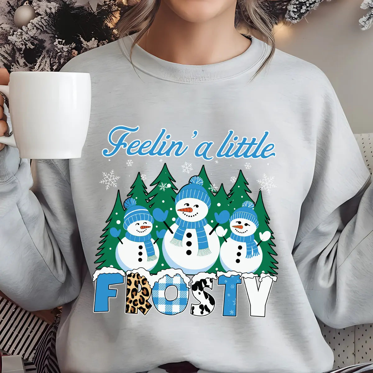 Light gray sweatshirt with “Feelin’ a Little Frosty” DTF transfer featuring three smiling snowmen, pine trees, snowflakes, and bold patterned lettering, perfect for winter apparel.